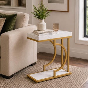 Könnte beinhalten: Ein Beistelltisch mit Marmor-Effekt-Platte und einem goldfarbenen Metallrahmen. Der Tisch hat eine rechteckige Ober- und Unterseite mit einem geschwungenen Design an den Seiten. Oben steht eine Vase mit Grünzeug.
