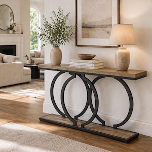 Puede incluir: Una consola de madera con una base de metal negro con tres diseños circulares. La encimera es de color marrón claro y la base tiene un estante inferior. Una lámpara, un jarrón y libros se exhiben en la mesa.