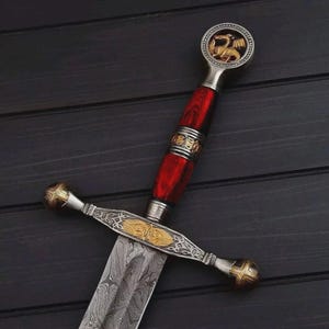 May include: A decorative dagger featuring a patterned Damascus steel blade, a vibrant red handle with metallic accents, and a crossguard with gold-toned engravings. The pommel is circular and displays a gold-coloured dragon emblem in the centre.
