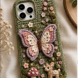 May include: A sage green crochet phone case featuring a large, dimensional butterfly with pink, cream, and lavender wings. The case is decorated with small embroidered pink flowers, a white-spotted red mushroom, and a small beige fawn figure.