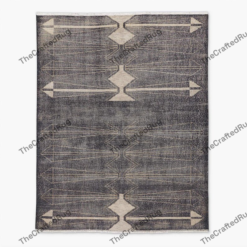 May include: A rectangular, dark gray rug with a geometric pattern. The design features diamond and arrow shapes in a lighter beige color, creating a contrasting visual effect. The rug has a fringe along the top edge, adding a textured detail.