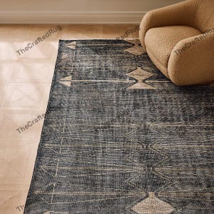 May include: A dark blue area rug with a geometric pattern of beige arrows and diamond shapes. The rug has a distressed, vintage look and is placed on a light wood floor. A beige armchair is in the background.