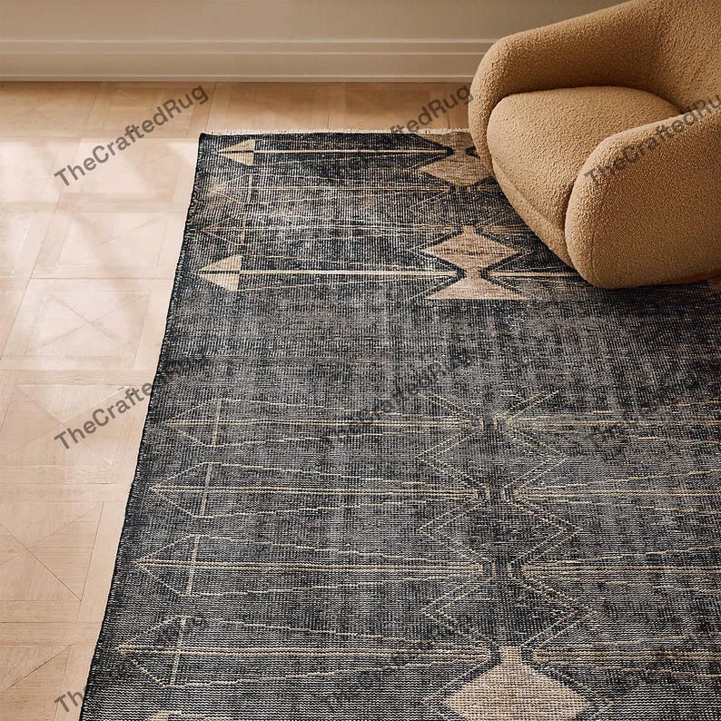May include: A dark blue area rug with a geometric pattern of beige arrows and diamond shapes. The rug has a distressed, vintage look and is placed on a light wood floor. A beige armchair is in the background.