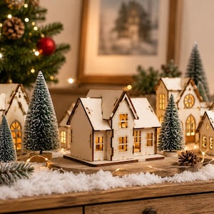 以下が含まれることがあります： 明るい色の壁と尖った屋根を持つ木製のミニチュアハウスのコレクションで、内側から照らされた窓の切り抜きが特徴です。家々は、雪のような表面の上に置かれた小さな緑のボトルブラシツリーや松ぼっくりの間に配置されています。