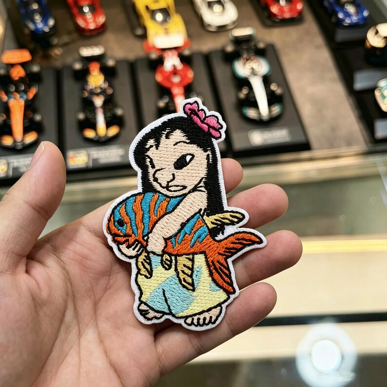 May include: An embroidered patch featuring a cartoon character with black hair and a pink flower, holding a large orange fish with blue stripes. The character wears a light blue and yellow patterned garment, all outlined in white thread.
