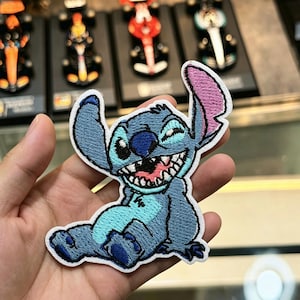 May include: An embroidered patch featuring the character Stitch in a seated position. The design displays a blue creature with a wide, toothy grin, large ears with pink inner detailing, and a dark blue nose, all outlined with a white border.