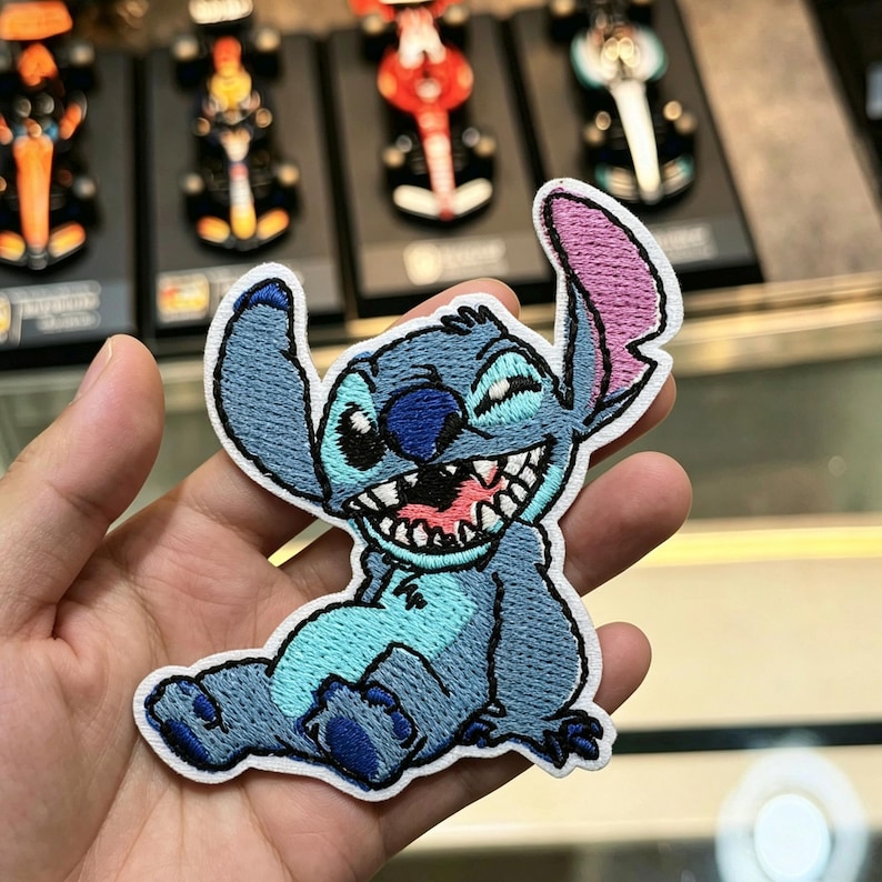 May include: An embroidered patch featuring the character Stitch in a seated position. The design displays a blue creature with a wide, toothy grin, large ears with pink inner detailing, and a dark blue nose, all outlined with a white border.