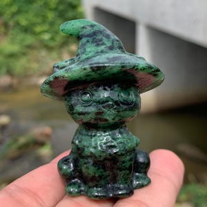 May include: A carved stone figurine of a cat wearing a pointed witch hat. The sculpture is made of a dark green mineral with black speckled inclusions throughout, featuring a smooth, polished surface and detailed facial features including eyes.