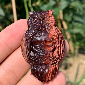 Peut inclure: Une sculpture en pierre œil-de-tigre brun rougeâtre représentant un hibou détaillé aux ailes déployées perché au-dessus d'un crâne humain. La pierre présente une texture fibreuse et chatoyante avec des tons acajou et cuivre profonds.