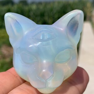 May include: A carved cat head figurine made of iridescent, semi-translucent opalite glass. The sculpture features pointed ears, almond-shaped eyes, and a small third eye symbol carved into the forehead between the ears, displaying a soft blue glow.