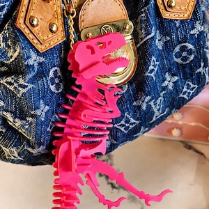 May include: A bright pink, articulated T-Rex dinosaur skeleton keychain hanging from a blue denim handbag. The handbag features a monogram pattern, brown leather trim, gold-tone metal hardware, and a central clasp with a circular logo emblem.
