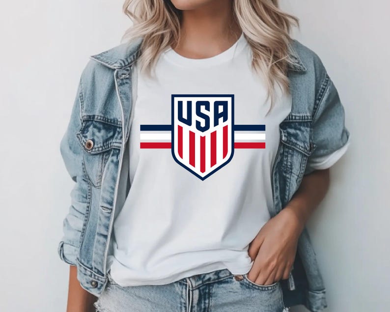 May include: White t-shirt featuring a USA shield logo with red, white, and blue stripes. The shirt has a crew neck and short sleeves. The graphic design is centered on the chest.