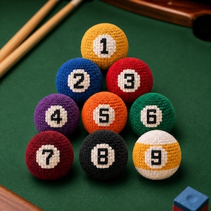 May include: A collection of colorful, crocheted billiard balls arranged on a green felt surface. Each ball is a different color and features a white number from 1 to 9. Two wooden pool cues are visible in the background.