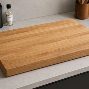 May include: A rectangular wooden cutting board with a natural light brown wood grain finish. The board features a thick, solid construction with smooth, rounded edges and a flat surface, showcasing the organic texture of the wood grain throughout.