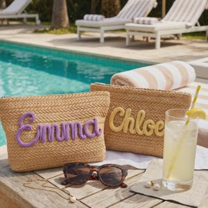 May include: Two woven straw zipper pouches, one with "Emma" in purple yarn and the other with "Chloe" in yellow yarn. Sunglasses, a necklace, earrings, and a drink with a lemon wedge are on a wooden table near a pool.