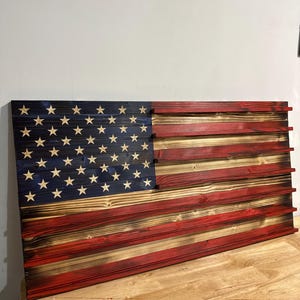 May include: Wooden American flag with a distressed finish. The flag features the traditional red and white stripes and a blue field with white stars. The wood grain is visible, adding to the rustic aesthetic. The flag is mounted on a wooden surface.
