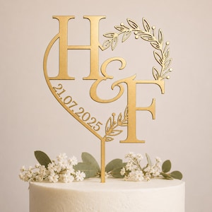 Op de afbeelding: Een goudkleurige taarttopper met de letters H en F gescheiden door een ampersand. Het ontwerp bevat een bladerkrans en de datum 21.07.2025 geïntegreerd aan de linkerkant. De topper heeft een enkele verticale prikker aan de onderkant.