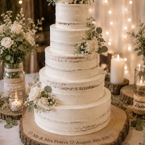 May include: A four-tier white semi-naked cake decorated with fresh white roses and greenery, resting on a round wooden slice base engraved with the text "Mr & Mrs Peters ♡ August 30th 2026 ♡". The cake features visible cake layers.