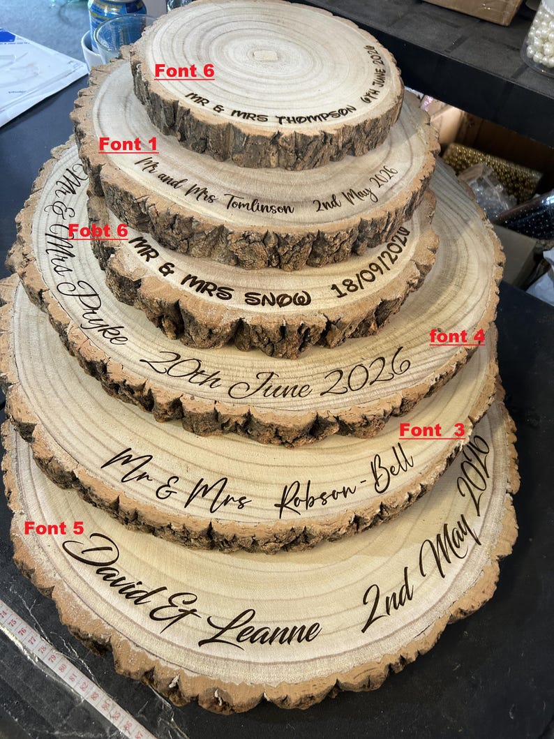 May include: A stack of five circular wooden tree slices with natural bark edges, each engraved with names and dates in various fonts labeled Font 1, 3, 4, 5, and 6. The engravings include Mr & Mrs Thompson, Tomlinson, Snow, Pyke, Robson-Bell, and David & Leanne.