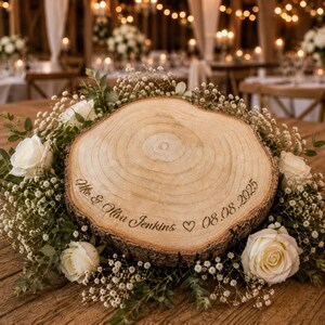 May include: A circular wood slice centerpiece featuring natural bark edges and a smooth, light-colored top surface. The wood is engraved with the text "Mike & Oliva Jenkins ♡ 08.08.2025" and is surrounded by a decorative wreath of white roses and baby's breath.