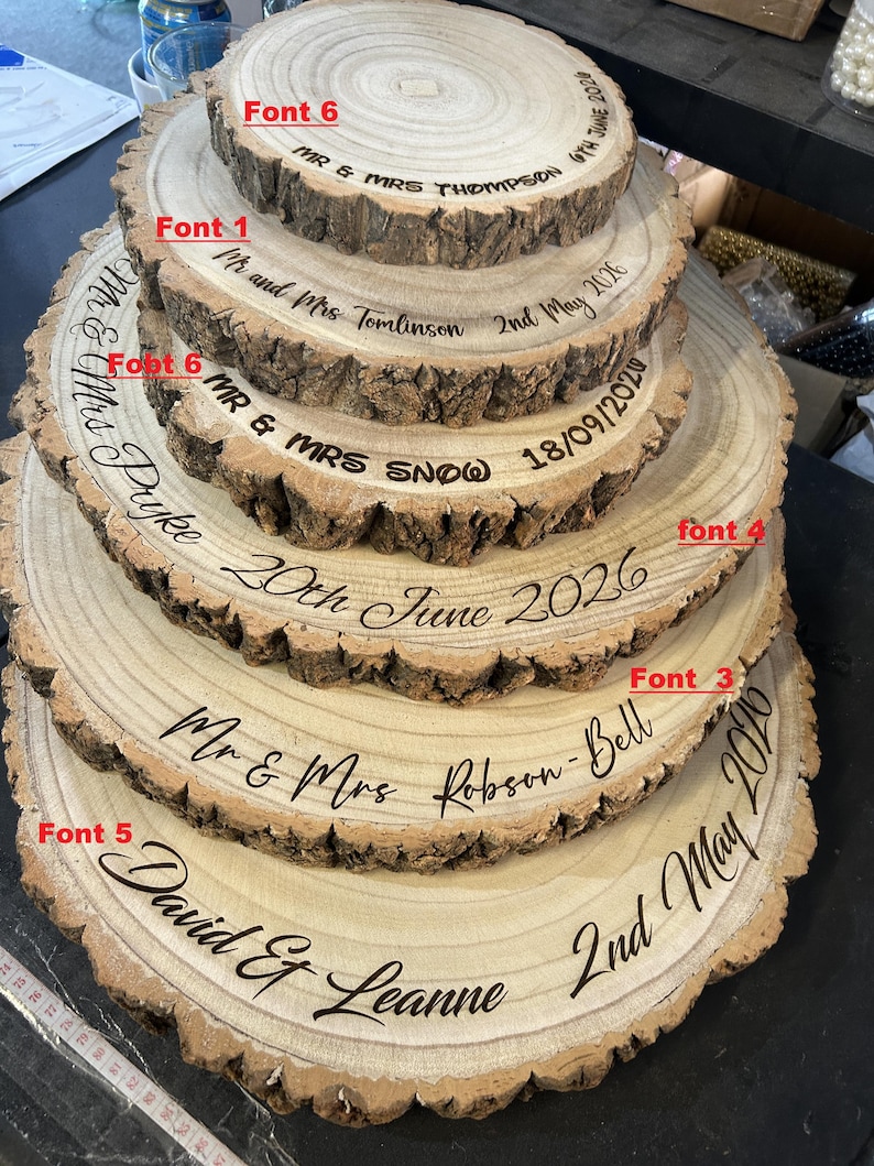 May include: A stack of five circular wood slices with natural bark edges, each engraved with names and dates in various fonts labeled Font 1, 3, 4, 5, and 6. The engravings include Mr & Mrs Thompson, Tomlinson, Snow, Pyke, Robson-Bell, and David & Leanne.