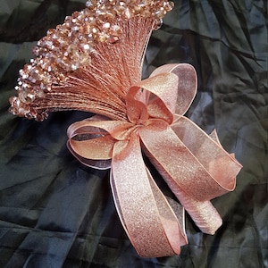 Puede incluir: Un ramo de novia de color oro rosa hecho de cuentas de cristal y alambre, atado con una cinta de brillo de oro rosa.