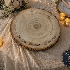 May include: A round wooden cake stand with natural bark edges, featuring the engraved text "Mr & Mrs Bartlet - 25.04.2026" with heart symbols. A circular label indicates an XL size suitable for 12-14 inch cakes.