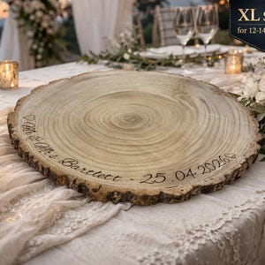 May include: A round wooden cake stand with natural bark edges, featuring the engraved text "Mr & Mrs Bartlett - 25.04.2026" and a heart symbol. A black label in the corner displays "XL SIZE for 12-14" cakes" in white text.
