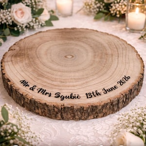 May include: A round wooden log slice cake stand with natural bark edges. The top surface is engraved with the text "Mr & Mrs Sgubič 13th June 2026" in a dark script font. The wood features visible grain patterns and a light brown color.
