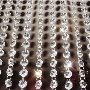 May include: A decorative curtain composed of clear, faceted crystal beads. The beads are strung in vertical lines, producing a shimmering, elegant effect. The background is a blurred, neutral tone.