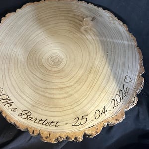 May include: A circular wooden slice with natural bark edges featuring a light tan wood grain surface. The wood is engraved with the text "Mr & Mrs Bartlett" and the date "25.04.2026" in a cursive script, accompanied by two small heart outlines.