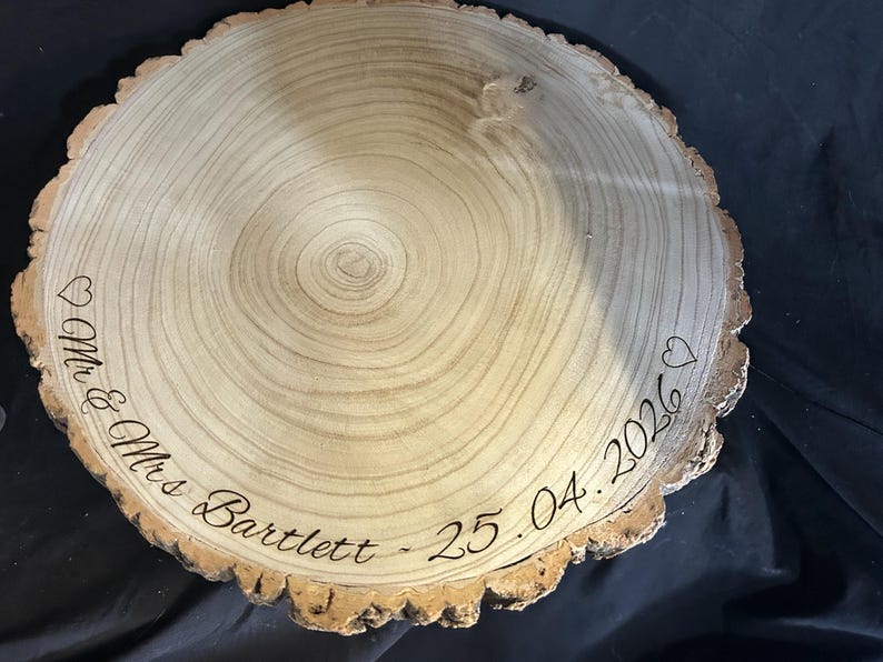 May include: A circular wooden slice with natural bark edges featuring a light tan wood grain surface. The wood is engraved with the text "Mr & Mrs Bartlett" and the date "25.04.2026" in a cursive script, accompanied by two small heart outlines.