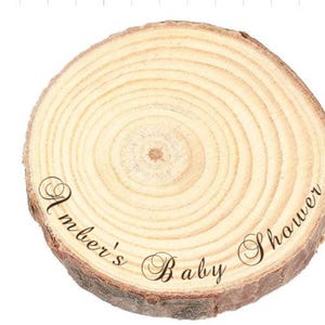 May include: A circular wooden slice with natural bark edges and visible tree rings. The surface features the black cursive text Amber's Baby Shower printed across the center, showcasing the light tan wood grain and organic texture of the wood.