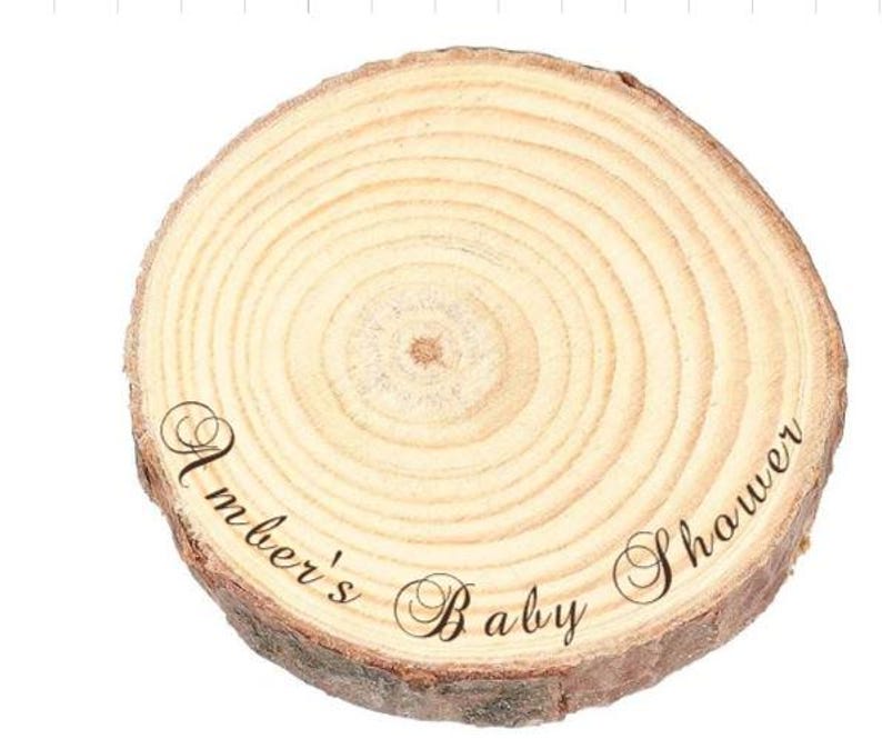 May include: A circular wooden slice with natural bark edges and visible tree rings. The surface features the black cursive text Amber's Baby Shower printed across the center, showcasing the light tan wood grain and organic texture of the wood.