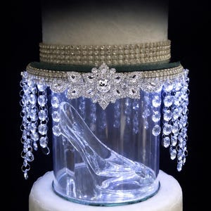 May include: A clear glass slipper inside a cylindrical display case, topped with a decorative band of pearls and hanging crystal beads. The text reads CRYSTAL GLASS SLIPPER Elegant Center Display. The display sits atop a white tiered base.