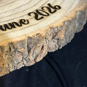 May include: A circular wooden slice with natural bark edges featuring the laser-engraved text "June 2026" on its light-colored, grained surface. The wood shows visible growth rings and a rough, textured bark perimeter in shades of brown and tan.