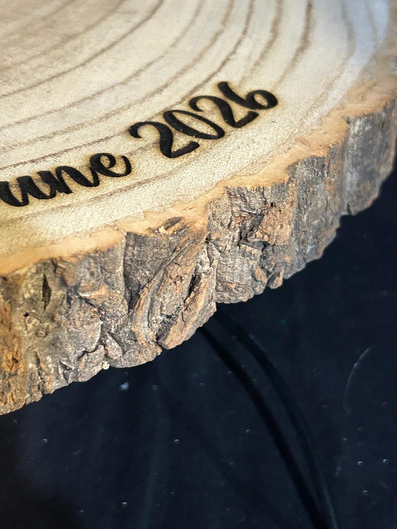 May include: A circular wooden slice with natural bark edges featuring the laser-engraved text "June 2026" on its light-colored, grained surface. The wood shows visible growth rings and a rough, textured bark perimeter in shades of brown and tan.
