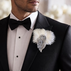 May include: A formal black tuxedo with a white dress shirt and black bow tie. A decorative brooch with white feathers and sparkling crystals is pinned to the lapel, adding a touch of elegance to the ensemble.