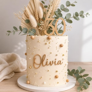May include: A cylindrical cake with smooth cream-coloured frosting, decorated with gold and white spherical sprinkles. The cake features a wooden script topper reading Olivia and a wooden number 30 topper, accented by dried palm leaves and eucalyptus.