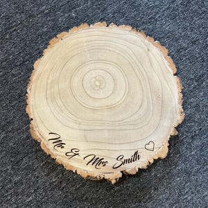 May include: A circular wooden slice with natural bark edges and visible tree rings. The surface is engraved with the black text "Mr & Mrs Smith" followed by a small heart outline. The wood features light tan tones with a smooth, sanded finish.