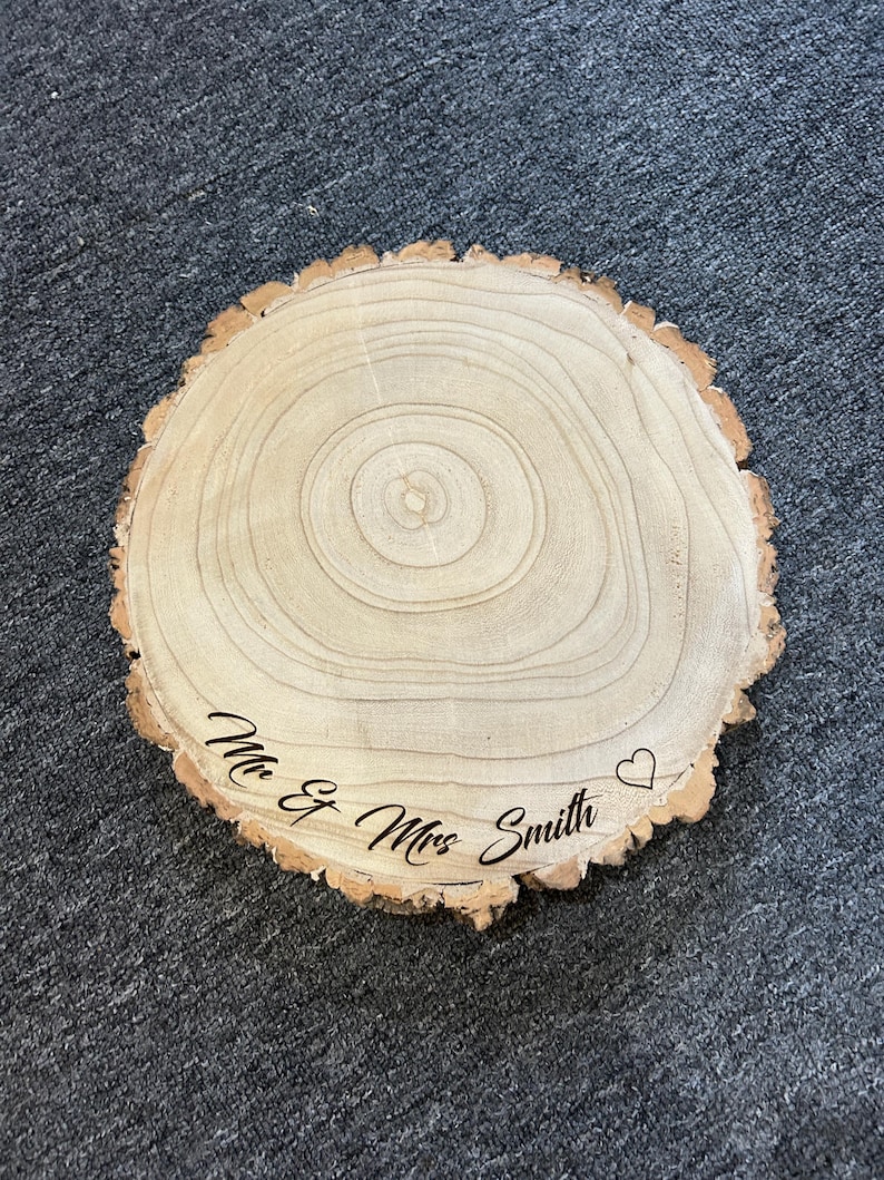 May include: A circular wooden slice with natural bark edges and visible tree rings. The surface is engraved with the black text "Mr & Mrs Smith" followed by a small heart outline. The wood features light tan tones with a smooth, sanded finish.