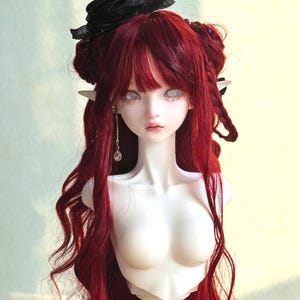 May include: A porcelain doll with long, flowing red hair and pale skin. The doll has a delicate face with large, blank eyes and a small mouth. It wears a black floral headpiece and a dangling earring. The doll's torso is visible, and it has pointed ears.