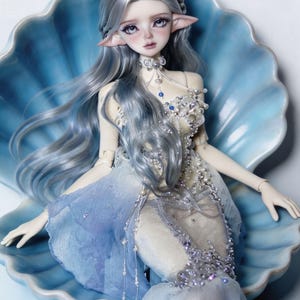 May include: A fantasy doll with long, silver hair and pointed ears, dressed in a flowing blue and white gown, adorned with beads. The doll is posed inside a large, blue seashell, creating a whimsical and ethereal scene.