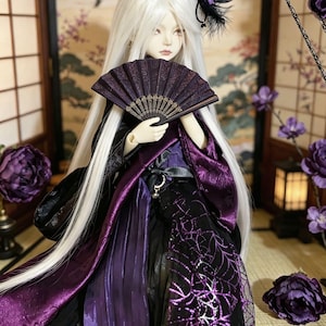 Pode incluir: Uma boneca com cabelo comprido branco e um leque roxo. A boneca usa uma roupa preta e roxa com um desenho de teia de aranha. Uma pena preta e uma flor roxa adornam a cabeça da boneca. O fundo inclui uma tela japonesa e flores roxas.