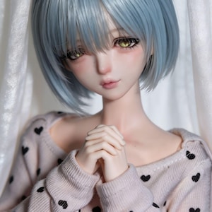 Könnte beinhalten: Eine Gelenkpuppe mit kurzem, hellblauem Haar und leuchtend gelben Augen. Die Puppe hat einen hellen Teint und trägt einen langärmeligen, hellrosa Pullover mit einem sich wiederholenden Muster aus kleinen, schwarzen, gestickten Herzen.