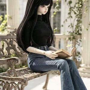 May include: A ball-jointed doll with long, straight black hair and bangs, wearing a black long-sleeved mock-neck top and blue denim jeans. The doll is seated and holding an open book with light-colored pages, displaying a neutral facial expression.