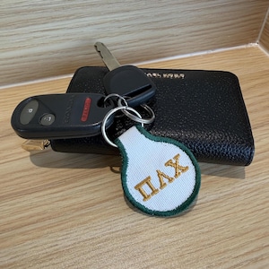 May include: A black leather wallet with a textured surface, a metal key, a black plastic car key fob with a red panic button, and a round white embroidered keychain featuring a dark green border and the gold Greek letters Pi, Lambda, and Chi.
