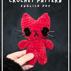 May include: A red crochet plush toy shaped like a cat with two black button eyes, a black stitched X for a mouth, and four thin metal whiskers on each side. The text CROCHET PATTERN ENGLISH PDF is displayed at the top of the image.