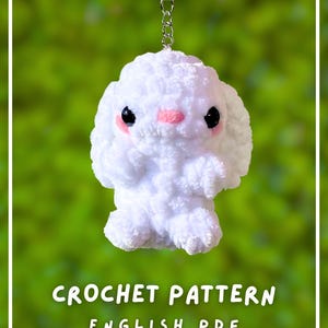 Op de afbeelding: Een witte gehaakte konijnen-sleutelhanger met zwarte ogen en roze wangen, bevestigd aan een zilveren metalen ring. De tekst CROCHET PATTERN ENGLISH PDF staat in het wit onderaan de afbeelding tegen een wazige groene achtergrond.