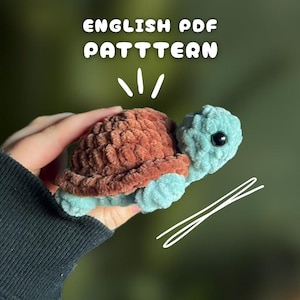 May include: A crocheted turtle with a brown shell and a light blue head and limbs. The turtle has a black eye and is held in a hand. The text "ENGLISH PDF PATTERN" is at the top of the image.